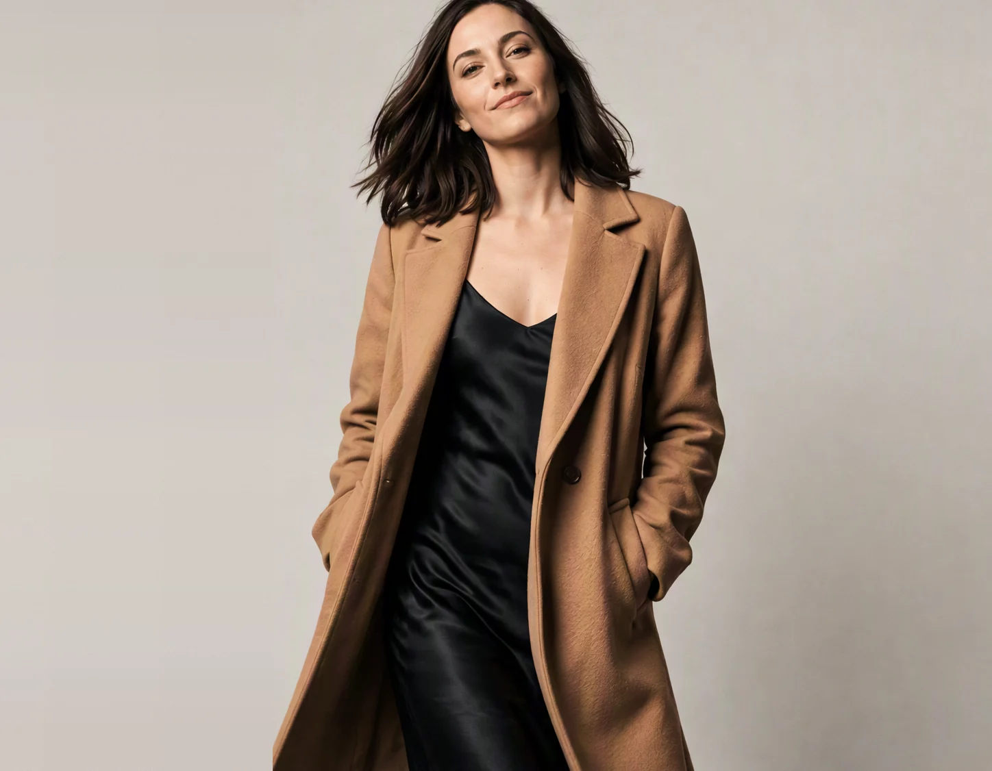 A woman wearing a camel coat over a black slip dress, photographed against a bone wall.